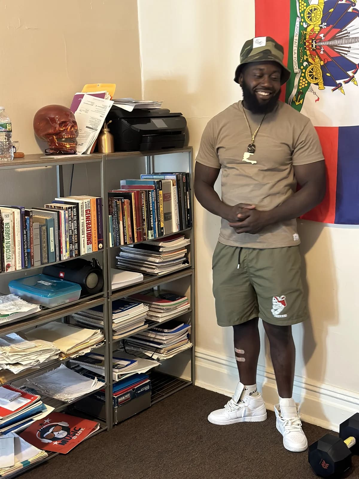Peeterson standing next to a full bookshelf at the XIISEEDS community space, Haitian flag in the background