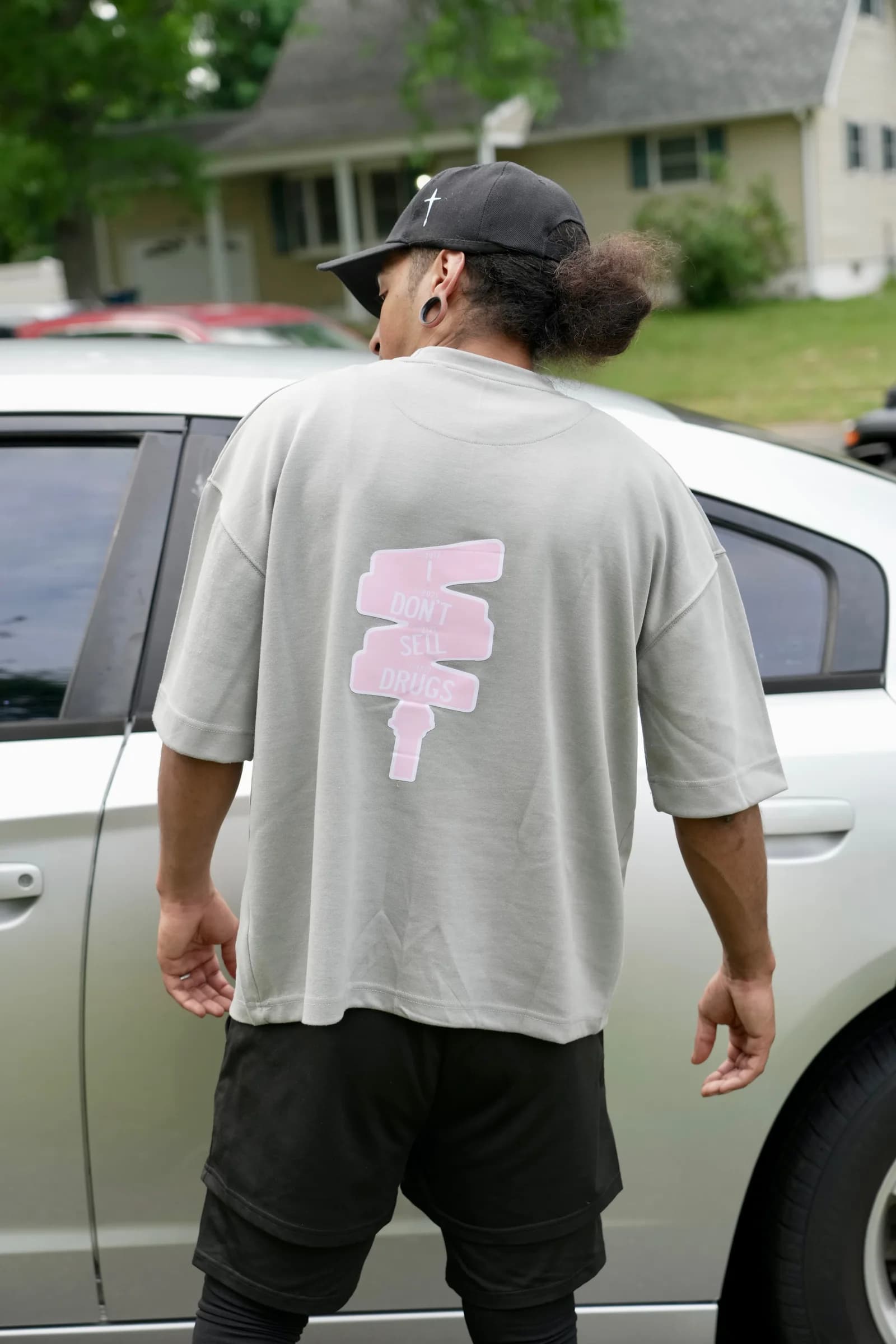 Man in gray XII SEEDS tee and cap with pink 'I Don't Sell Drugs' back graphic, standing by a car